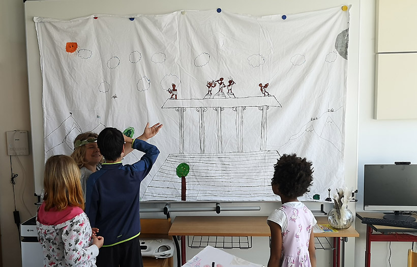 Vor einem selbstgestateten Wandbehang erklärt ein Kind seinen Mitschüler:innen etwas über die Antike.