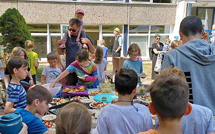 Kinder und Erwachsene stehen um ein großes Bufett voller Lebensmittel während des Schulfestes der DPFA-Regenbogen-Grundschule Liepzig.