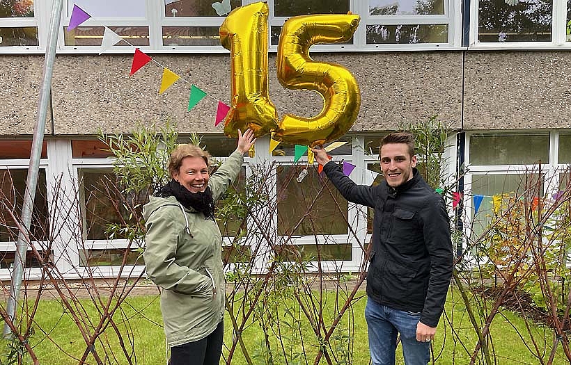 Vor einem Balloon "15" stehen eine Frau und ein Mann. Sie freuen sich über das 15. Jubiläum der DPFA Einrichtung Grundschule in Lei