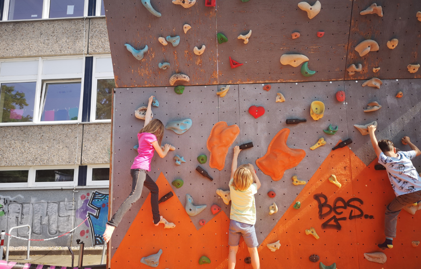 Drei Kinder kletten eine orange farbene Boulderwand hinauf.