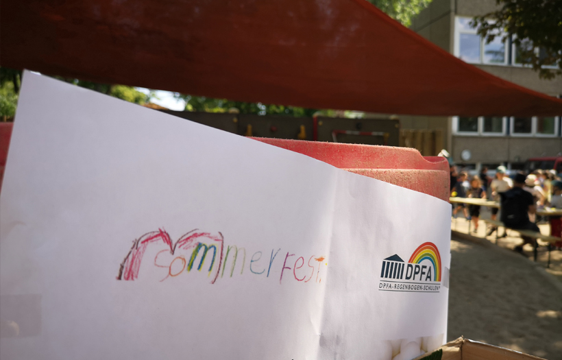 Auf einem weißen Blatt Papier hat ein Kind "Sommerfest" mit einem Regenbogen gemalt.