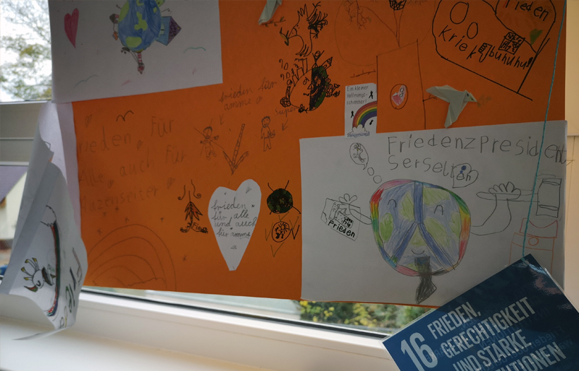 Ein orangenes Plakat hängt an einem Fenster. Auf dem Plakat hat ein Schulkind über Frieden auf der Welt geschrieben.